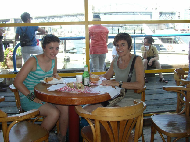 Zwei Frauen sitzen an einem Tisch mit Snacks in einem Schiff, mit anderen Menschen im Hintergrund, die die Aussicht genießen, einem anderen Schiff, einer Pflanze, einer Brücke, einem Himmel, einem Swimmingpool und Gebäuden im Hintergrund.