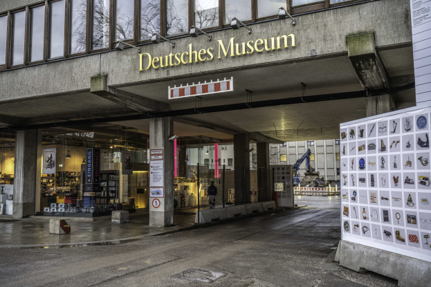 Außenansicht des Deutschen Museums in Berlin, das ein großes Glasgebäude mit Säulen, ein Namensschild und eine informative Anzeige auf der rechten Seite sowie Bäume und andere Gebäude im Hintergrund zeigt.