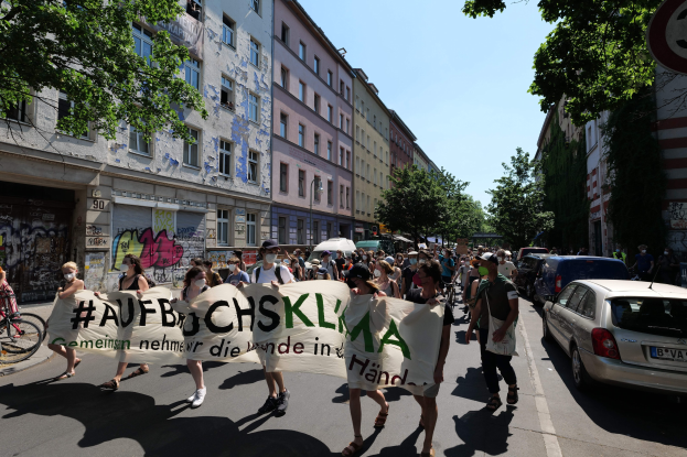 Eine Gruppe von Menschen marschiert auf einer Straße in Berlin, hält ein Transparent, mit geparkten Autos auf der rechten Seite, einem Radfahrer auf der linken Seite und Gebäuden, Bäumen und einem Schild im Hintergrund unter einem sichtbaren Himmel.