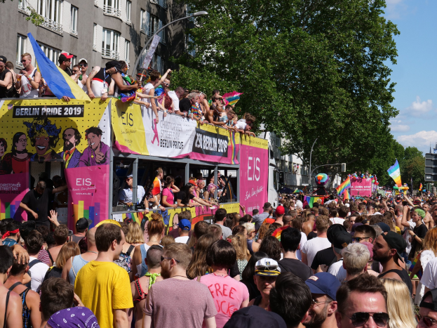 Eine große Menge marschiert während des Christopher Street Day 2019 in Berlin die Straße entlang, viele tragen Mützen und Schutzbrillen, einige halten Fahnen, mit Bannern, Laternenmasten, Bäumen und Gebäuden im Hintergrund unter einem bewölkten Himmel.