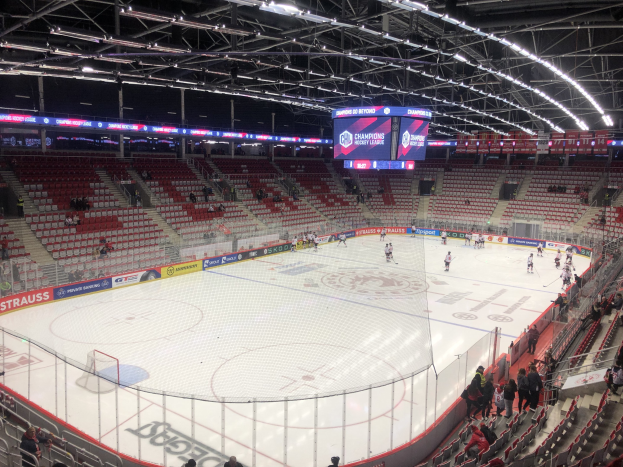 Eishockeyspiel in einer großen Arena mit Zuschauern, eine von einem Zaun umgebene Eisfläche, ein Display, das "St. Louis Blues vs Detroit Red Wings" anzeigt, Metallrahmen, Deckenleuchten und ein Dach.