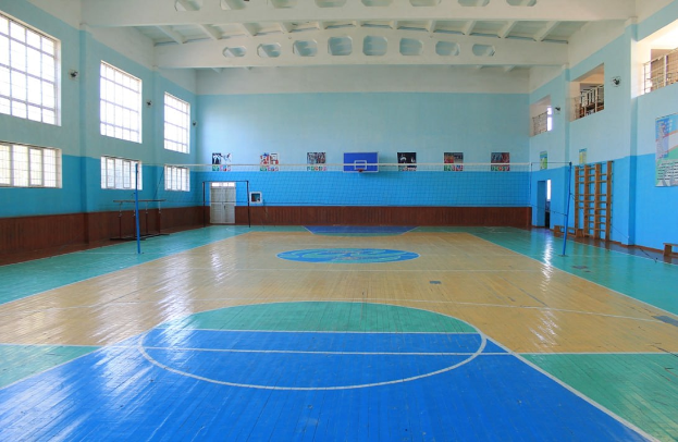 Großer Turnhalle mit einem blauen und weißen Basketballfeld, einem Netz mit Pfosten, einer Tür, Fenstern, Wandplakaten, einem Geländer und einem Dach.