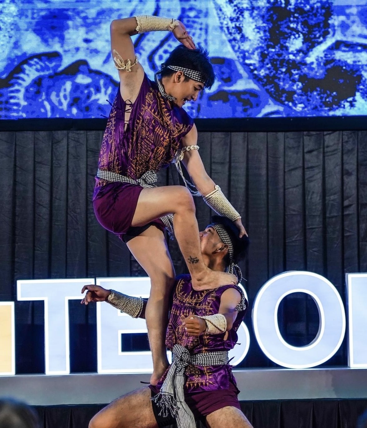 Zwei Performer in violetten Kleidern führen einen akrobatischen Hebeflug auf der Bühne vor einem Publikum aus, mit einem schwarzen Vorhang und einer Leinwand im Hintergrund.