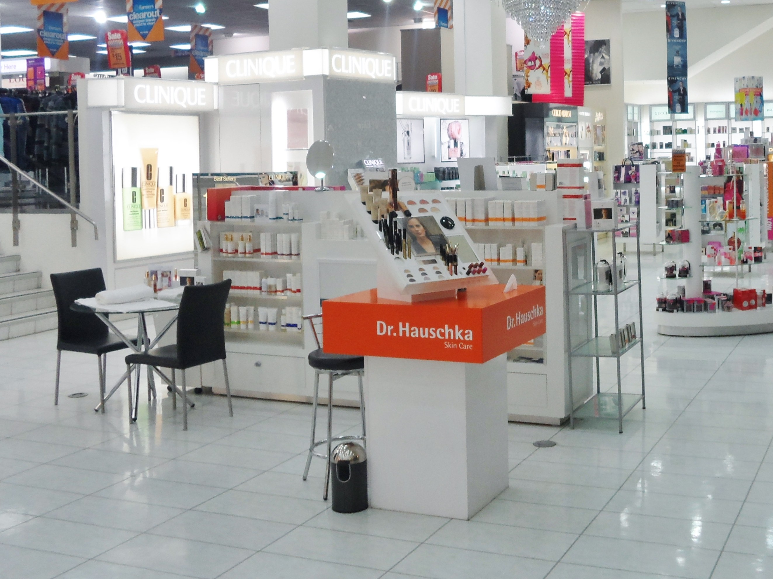 Ein Geschäft mit verschiedenen Kosmetikartikeln, Möbeln und Dekorationen, darunter Regale, Stühle, ein Tisch, ein Müllbehälter, eine Lampe, Schilder, eine Treppe, Kleidung und Deckenleuchten.