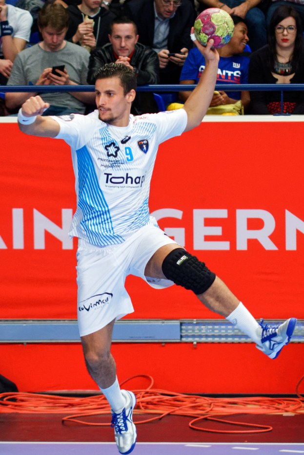 Ein Mann in der Luft, der einen Handball hält, mit einer Gruppe von Menschen und einem Banner mit Text im Hintergrund.