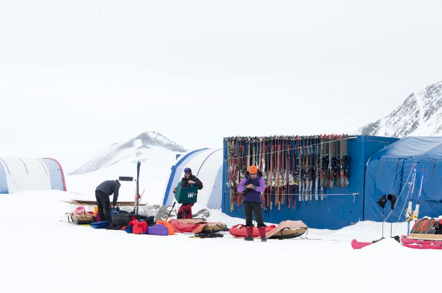 Drei Personen stehen auf einer schneebedeckten Landschaft mit verstreuten Taschen, Zelten mit Skiern im Hintergrund und schneebedeckten Hügeln unter einem klaren Himmel.