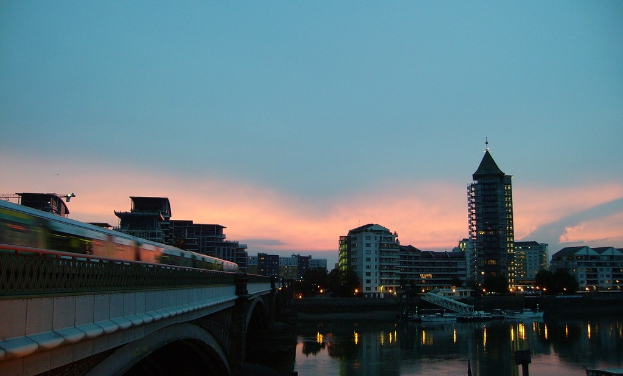 Ein Fluss mit weißen Booten auf dem Wasser, eine Brücke mit einem Zug auf der rechten Seite, Gebäude, Bäume, Lichter und Wolken im Hintergrund.