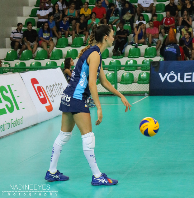 Frau im Volleyball in Aktion mit einem Ball in der Luft auf einem Feld mit Werbetafeln, Stufen, einer Wand und einer Gruppe von sitzenden Menschen im Hintergrund.