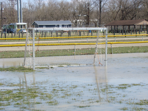 Ein Fußballtor steht in der Mitte eines überfluteten Feldes mit Gras und Wasser, umgeben von Hütten, Pfählen, Bäumen und Fahrzeugen unter einem klaren blauen Himmel.