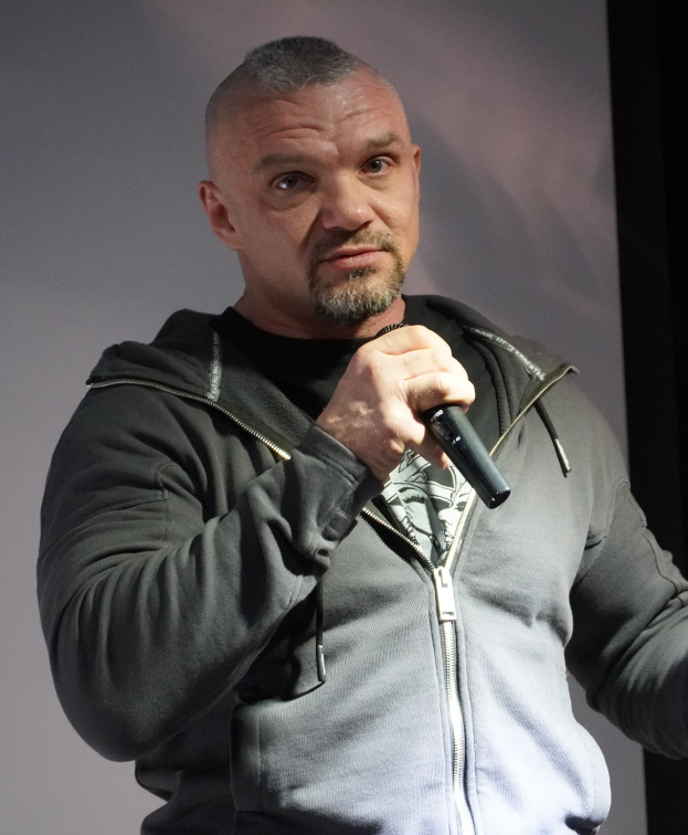 Comedian in black suit and white shirt, holding a microphone, with slicked-back hair and a confident expression, standing in front of a wall.