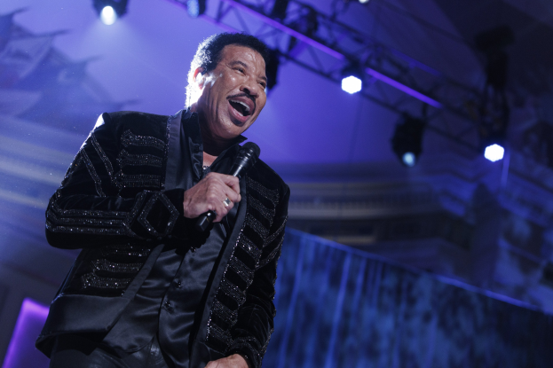 Lionel Richie performt bei den Grammys, trägt ein schwarzes Kleid und hält ein Mikrofon auf einer hell erleuchteten Bühne.