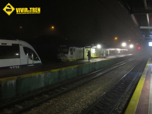 Eine BahnhofsSzene mit zwei Zügen, einem auf der linken und einem in der Mitte, mit Menschen auf einem Fußweg zwischen ihnen, Pfählen und Lampen im gesamten Bild.