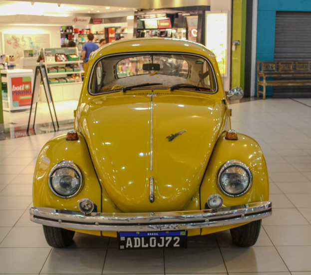 Ein gelber Volkswagen Käfer parkt auf einem Einkaufszentrum, mit verschiedenen Objekten, Menschen und Beschriftungen im Hintergrund.