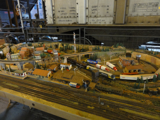 Eine Miniaturszene auf einem Tisch mit einer Modelleisenbahnschiene, einem Zug, Gebäuden, Fahrzeugen, einer Brücke, einem Turm und Bäumen.