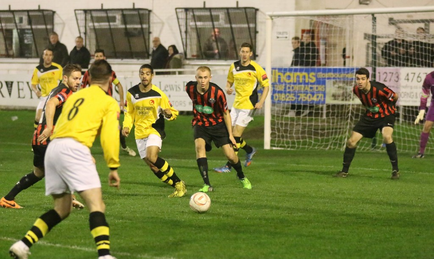 Ein Sportplatz mit mehreren Männern, die Fußball spielen, ein Netz auf der rechten Seite, eine Tafel mit Text im Hintergrund und Zuschauern, die hinter der Tafel stehen, sowie eine Wand und Fenster weiter im Hintergrund.