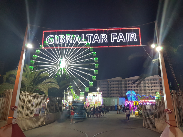 Der Eingang zur Gibraltar-Messe bei Nacht, beleuchtet von einem großen beleuchteten Riesenrad und einer leuchtenden Namensplakette, mit Menschenmassen, Bäumen und Gebäuden im Hintergrund unter einem dunklen Himmel.