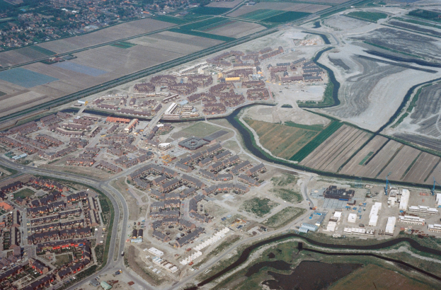 Luftaufnahme einer Stadt mit vielen Gebäuden, Straßen, Feldern, Bäumen und einem zentralen Reservoir, umgeben von städtischen und natürlichen Bereichen.