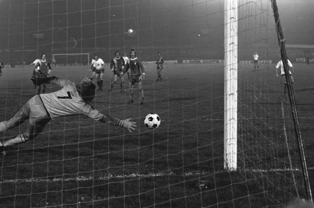 Ein Schwarz-Weiß-Foto eines Fußballspielers, der vor einem Tor einen Ball hält, umgeben von einer Gruppe von Menschen auf dem Boden. Das Torpfosten ist auf der rechten Seite des Bildes zu sehen, im Hintergrund gibt es Werbetafeln und Lichter.