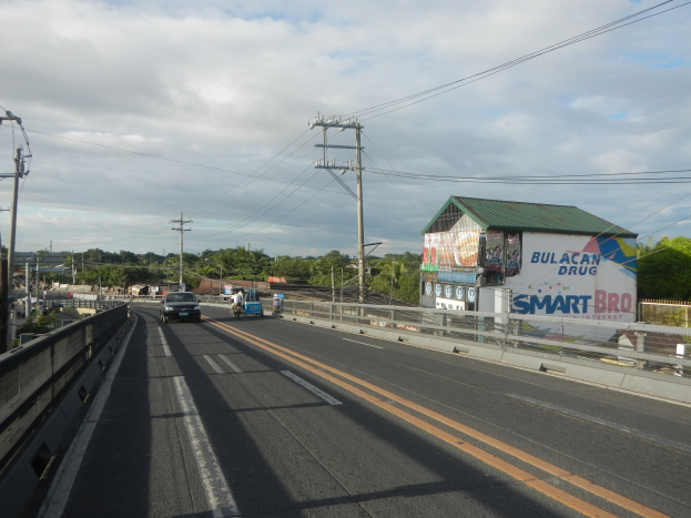 Eine Fahrbahn mit Fahrzeugen, Strommasten mit Kabeln, ein Schuppen mit einem Banner, Bäume, Gebäude, ein bewölkter Himmel und ein Straßenrand-Schild.
