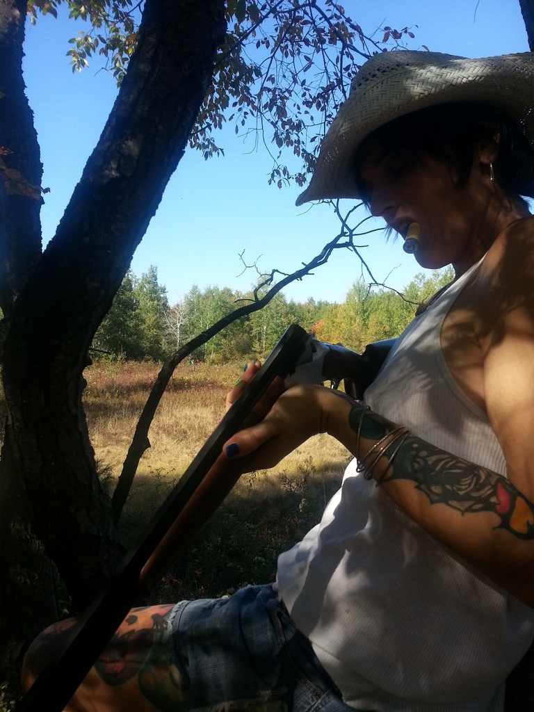 Eine Frau steht in der Mitte und hält eine Waffe und eine Zigarette im Mund, mit Bäumen im Hintergrund, Gras unten und Himmel oben.