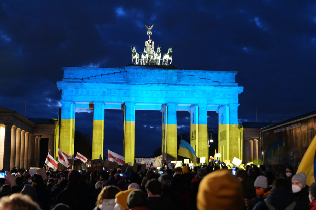 Eine Menschenmenge mit Fahnen und Plakaten steht vor dem Brandenburger Tor in Berlin, Deutschland, mit einer Banner auf der rechten Seite und den Torstatuen sichtbar unter einem bewölkten Himmel.