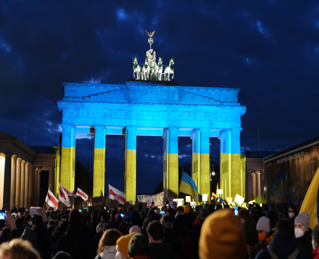 Eine Menschenmenge mit Fahnen und Plakaten steht vor dem Brandenburger Tor in Berlin, Deutschland, mit einer Banner auf der rechten Seite und den Torstatuen sichtbar unter einem bewölkten Himmel.