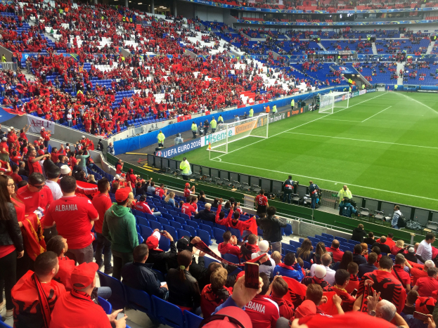 Eine große Menschenmenge in einem Stadion schaut einem Fußballspiel zu, mit Menschen, die auf Stühlen sitzen und stehen, Bannern, einem Torpfosten und Deckenleuchten.