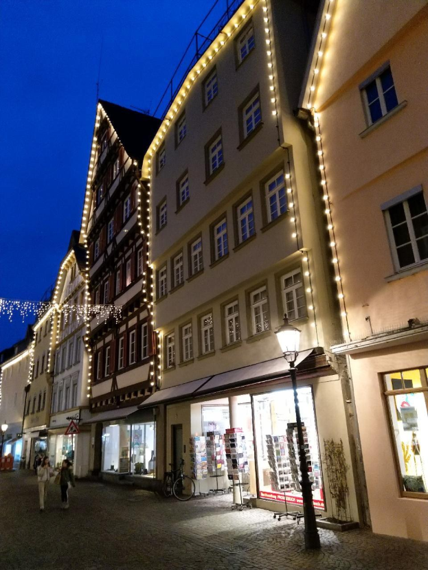 Headline in Rothenburg ob der Tauber, Germany, decorated with Christmas lights, with people walking and a bicycle parked nearby, flanked by buildings with windows under a visible sky.