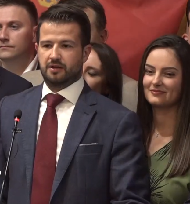 Ein Mann im Anzug und mit Krawatte steht an einem Rednerpult mit einem Mikrofon und spricht vor einer Menge mit einem Banner im Hintergrund, möglicherweise als Reaktion auf ein Regierungverbot von Social Media.
