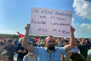 Ein Mann hält ein Schild mit der Aufschrift "Aserbaidschanische Protestler demonstrieren in Ankara für den Rücktritt von Präsident Ilham Aliyev" vor einer großen Menge, einige mit Fahnen und Handys, unter einem blauen und weißen Himmel mit Bäumen im Hintergrund.