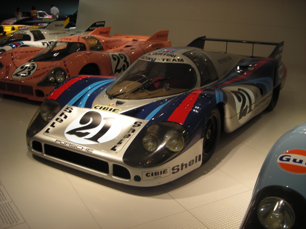Ein Porsche 917K Le Mans-Rennwagen in einem Museum neben anderen Sportwagen ausgestellt, mit Menschen, die zuschauen, und einer Wand im Hintergrund, mit einer Texttafel auf der linken Seite.