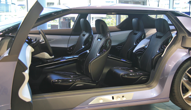 Das Innenleben eines Autos mit geöffneter Tür, das luxuriöse Ledersitze, Lenkrad und Räder zeigt, mit Gebäuden und Bäumen im Hintergrund.