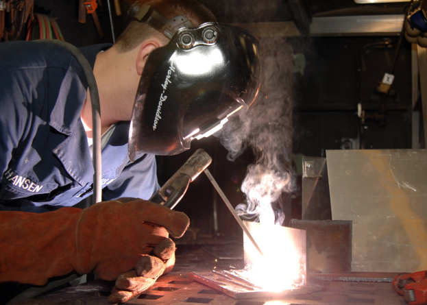 Ein Schweißer in Schutzausrüstung bedient eine Schweißmaschine und erzeugt dabei Rauch, während er an einem Metallstück arbeitet, wobei verschiedene Werkzeuge und Gegenstände im Hintergrund zu sehen sind.