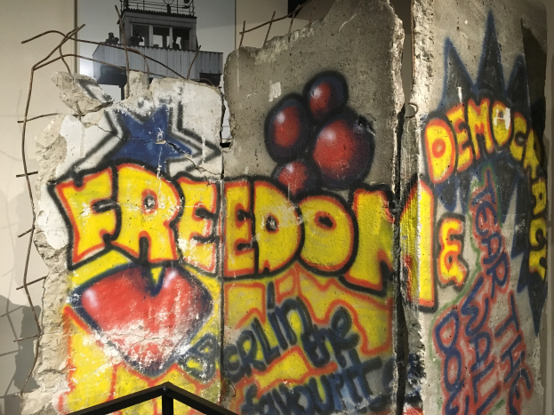 Mauer mit Graffiti, das in fetten schwarzen Buchstaben 'Freiheit und Demokratie' liest, ein Geländer unten und ein Gebäude mit Fenstern im Hintergrund.