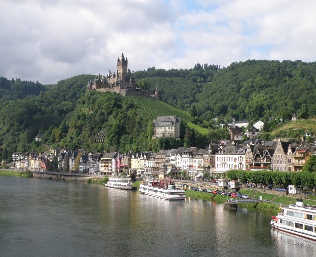 Ein malerischer Blick auf den Rhein in Deutschland mit einer Burg auf einem Hügel im Hintergrund, Booten auf dem Fluss, Fahrzeugen auf einer nähergelegenen Straße und einem bewölktem Himmel.