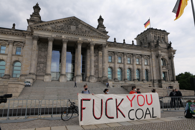 Eine Gruppe von Menschen hält ein Transparent mit der Aufschrift "Fuck You Coal" vor dem Reichstaggebäude in Berlin, Deutschland, das mit Säulen, Fenstern, Bögen und Statuen geschmückt ist und von einem Metallzaun umgeben ist. Es gibt Stufen, die zum Eingang führen, ein Fahrrad in der Nähe, Bäume, einen Fahnenmast mit einer wehenden Flagge und einen bewölkten Himmel im Hintergrund.