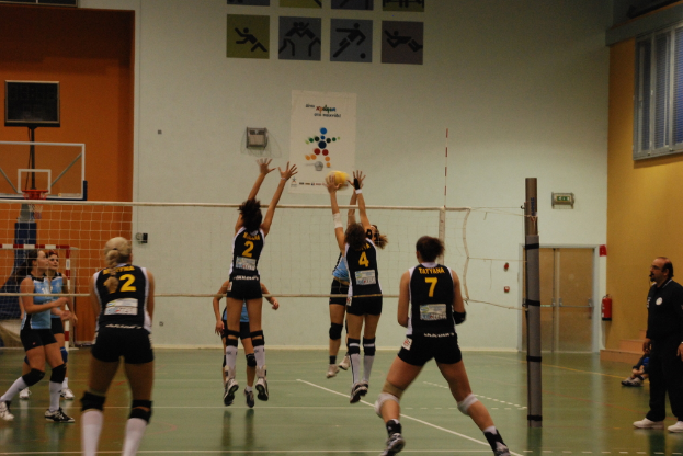 Ein Volleyballfeld mit spielenden Spielerinnen, mit einem Basketballkorb, Fenstern und einer Tür im Hintergrund.