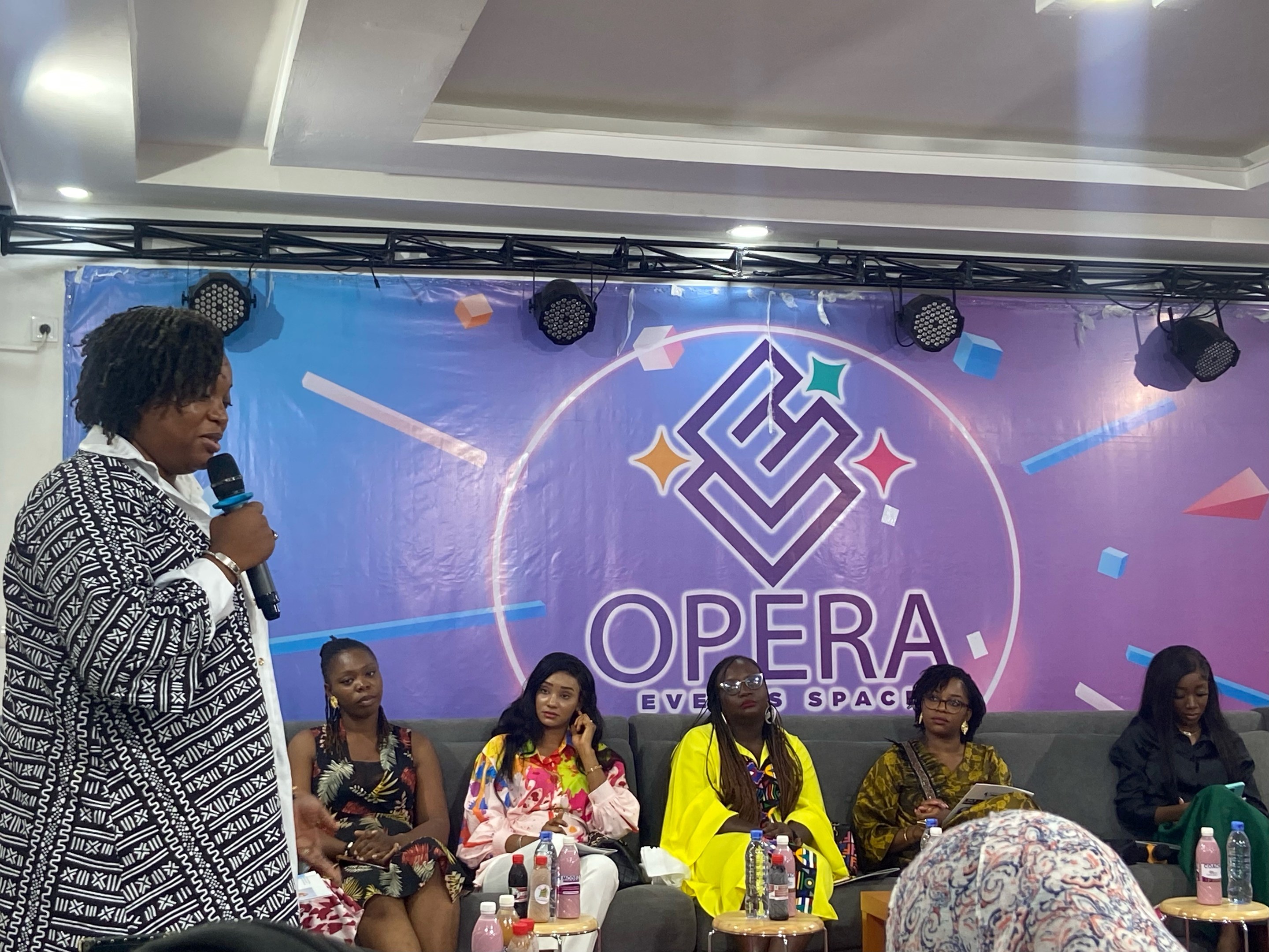 Frau steht vor sitzenden Frauen auf einer Couch, hält ein Mikrofon, mit Flaschen und Gegenständen auf den Tischen im Vordergrund, ein Banner mit Text im Hintergrund und Deckenbeleuchtung.