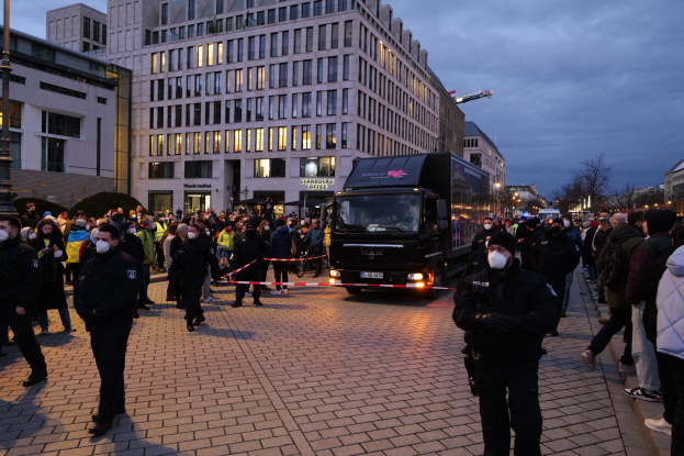 Eine Gruppe von Menschen steht in der Nähe eines Lastwagens auf einer Straße, die von Gebäuden, Bäumen und Laternen gesäumt ist, unter einem bewölkten Himmel, wobei einige eine Mütze und eine Maske tragen und ein Band an einem Pfahl im Vordergrund zu sehen ist.