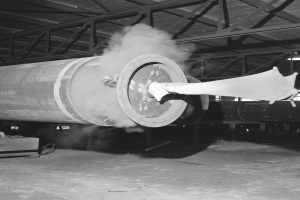 Ein Schwarz-Weiß-Foto einer großen Rohrleitung in einer Fabrik, aus der Rauch aufsteigt, mit einer Maschine links und anderen Gegenständen rechts, unter einem sichtbaren Fabriktor.