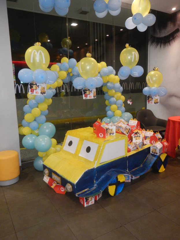 Eine McDonald's-Geburtstagsfeier mit einem gelben und blauen Lastwagen, der mit Ballons geschmückt ist, einem rot gedeckten Tisch mit Gegenständen, Hockern, einer Glaswand und Deckenlampen, die eine festliche Atmosphäre schaffen.