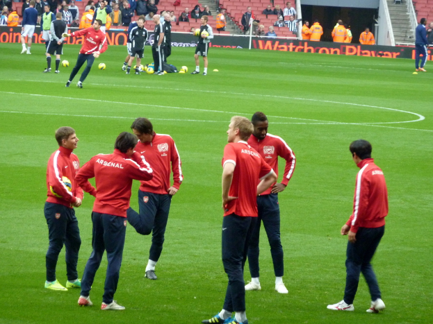 Eine Gruppe von Spielern auf einem grünen Feld mit Footballs und einer Zuschauerschaft im Hintergrund.