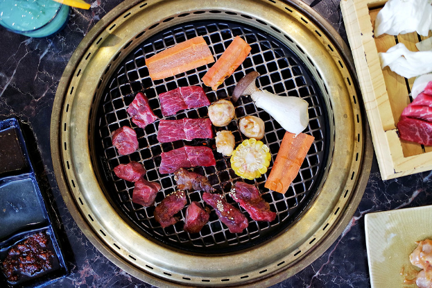 Ein Grill mit Fleisch, Mais, Karotten und Pilzen darauf, mit Tabletts und Tellern auf einem nahen Tisch.