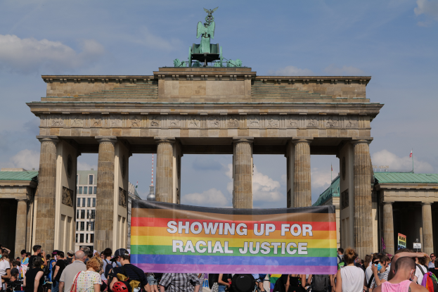 Eine Gruppe von Menschen steht vor dem Brandenburger Tor in Berlin, Deutschland, mit einer Tafel, auf der "Racial Justice" steht, vor den Säulen des Tors, einer Statue und Gebäuden im Hintergrund bei einem bewölkten Himmel.