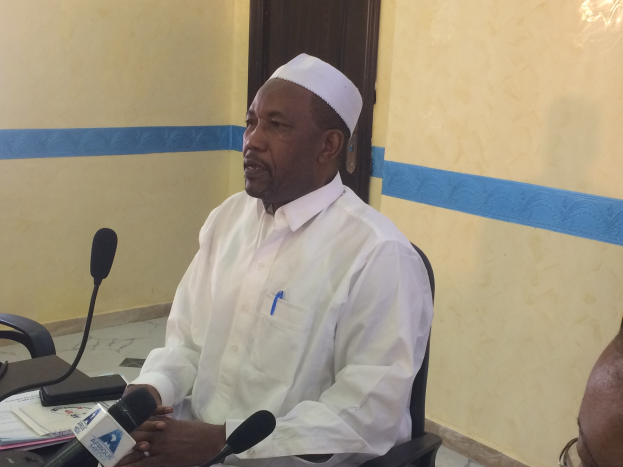 Ein Mann in weißem Hemd und Hut sitzt an einem Tisch mit Mikrofonen und spricht, während Papiere und Gegenstände auf dem Tisch zu sehen sind, und rechts von ihm sitzt eine Person mit Brille in einem Raum mit einer Wand und einer Tür im Hintergrund.