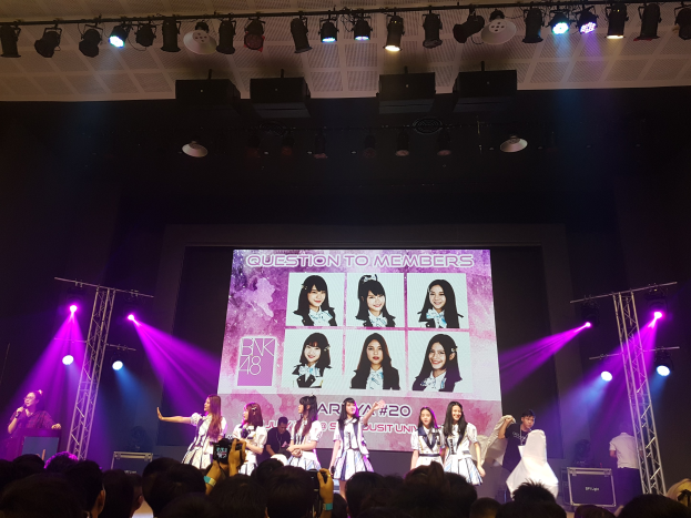 Eine Bühnenperformance mit BK48, bei der zwei Mitglieder auf der Bühne mit einem Mikrofon und einem Instrument stehen, umgeben von Trägern und hellen Lichtern, während die Menge unten aufzeichnet und ein großer Bildschirm Bilder der Gruppe anzeigt.