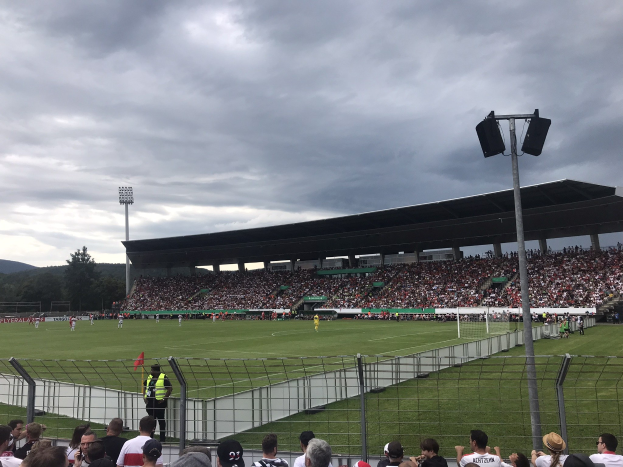 Eine große Menschenmenge schaut einem Fußballspiel in einem eingezäunten Stadion bei künstlichem Licht zu, mit Bäumen und Hügeln im Hintergrund unter einem bewölkten Himmel.