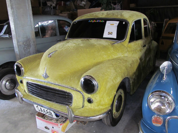 Mehrere Oldtimer vor einer Wand mit verschiedenen Zubehörteilen geparkt, mit einer Säule auf der linken Seite.