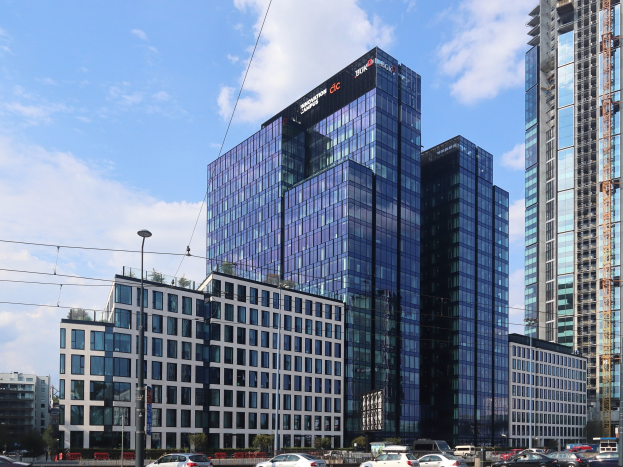 Außenansicht des Deutschen Bank-Hauptsitzes in Frankfurt, Deutschland, mit einem großen, mehrfenstrigen Gebäude, umgeben von städtischer Infrastruktur einschließlich Straßenlaternen, Fahrzeugen und Grünflächen.