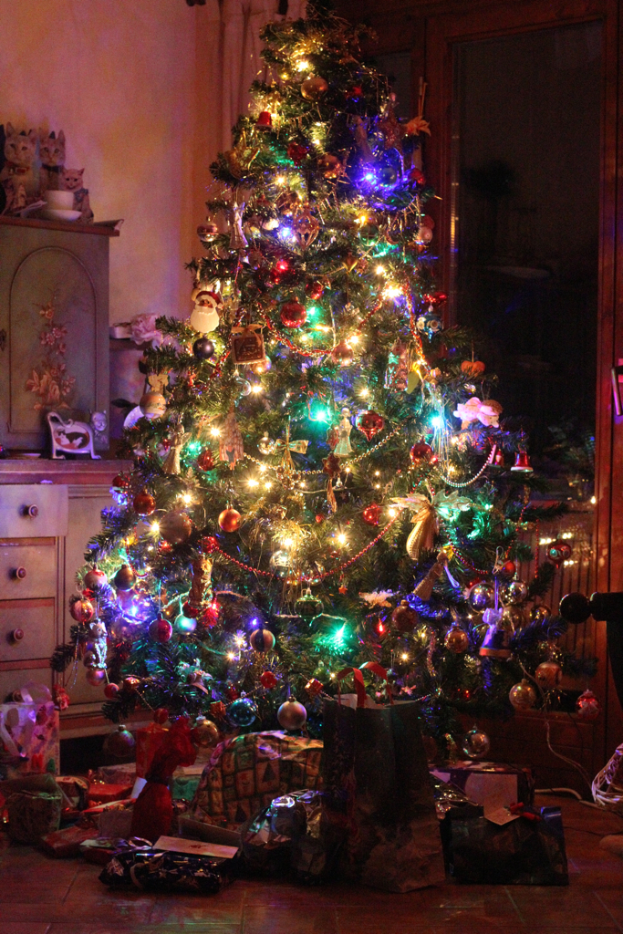 Ein Weihnachtsbaum mit Lichtern und Schmuck, Geschenke auf dem Boden, ein Schrank mit Gegenständen links und ein Fenster an der rechten Wand.